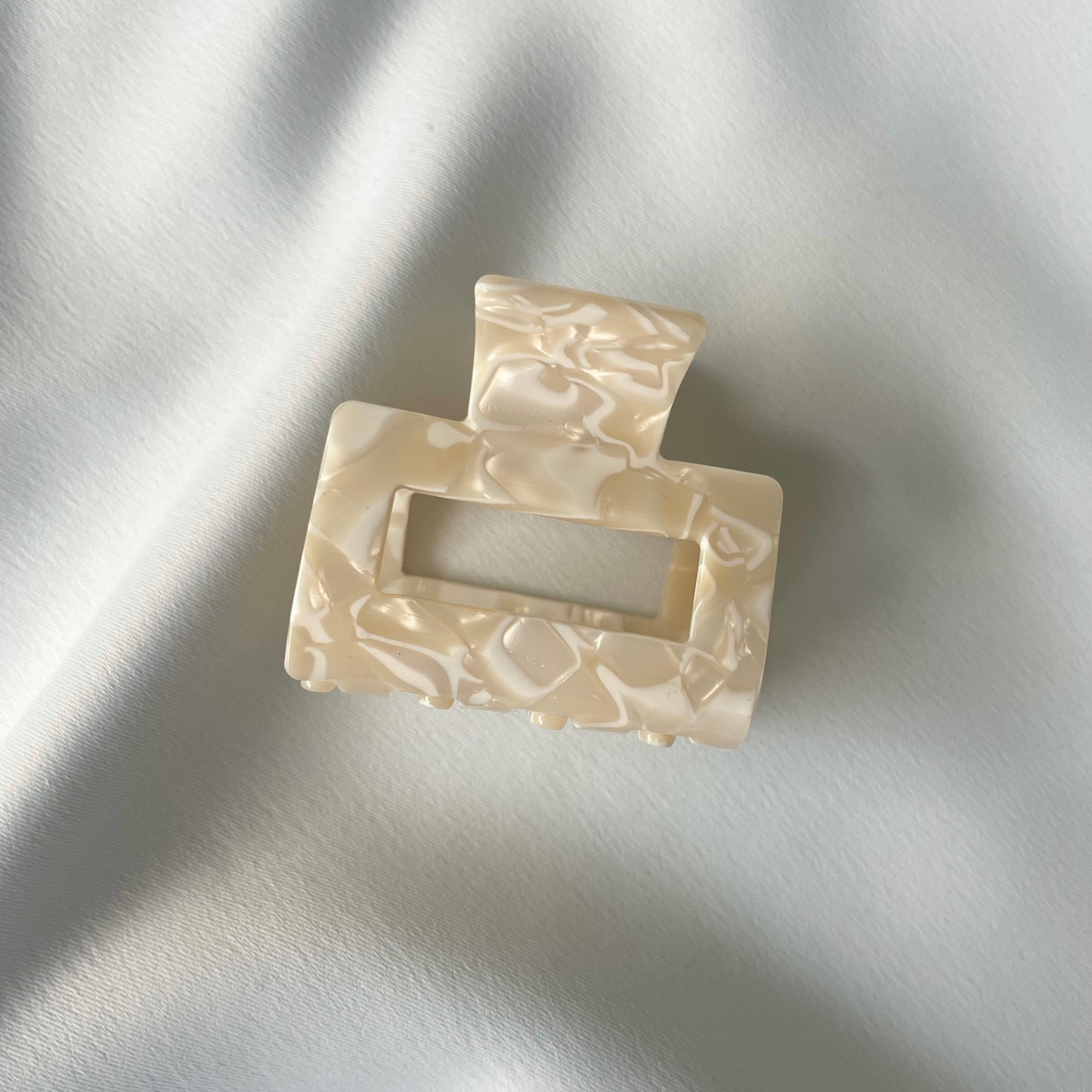 eine mittelgrosse quadratische Haarspange (5 cm) in beige mit weissen Details, die auf einem seidigen weissen Tuch liegt