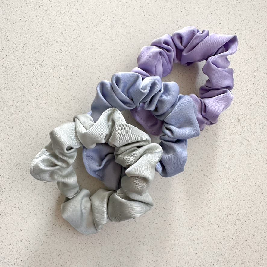Auf dem Bild sind drei Scrunchies in violett, hellblau & hellgrün zu sehen, die Scrunchies werden als Produktfoto auf einem neutralen Hintergrund präsentiert.