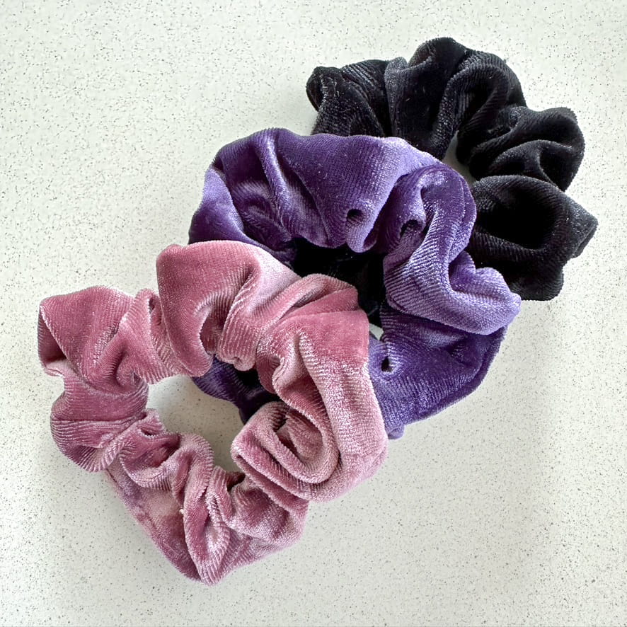 Auf dem Bild sind drei Scrunchies in rosa, violett & schwarz zu sehen, die Scrunchies werden als Produktfoto auf einem neutralen Hintergrund präsentiert.
